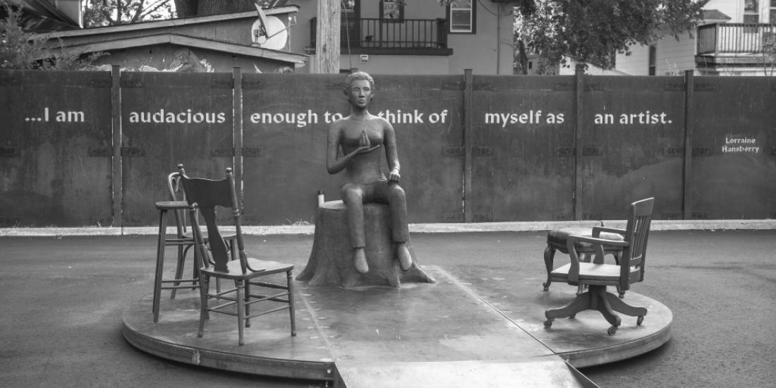 A sculpture of a seated person surrounded by 4 empty chairs. In the background is the text "I am audacious enough to think of myself as an artist," by Lorraine Hansberry.
