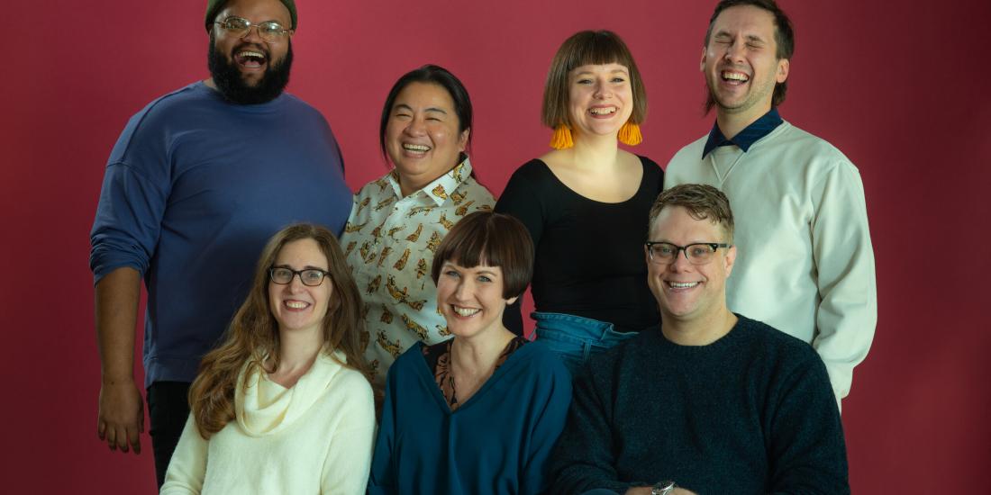 The 7 artistic directors of Red Eye smiling and laughing against a red backdrop.