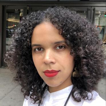 Joiri Minaya, a thirty-year-old Black woman artist slightly smiling while looking at the camera wearing a white shirt and red lipstick.