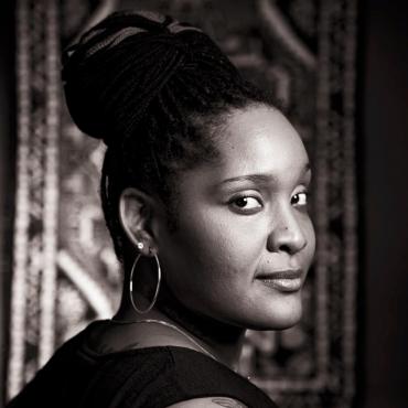 PaviElle, a Black woman artist and vocalist, posing in a headshot photograph, looking at the camera softly with a smile.