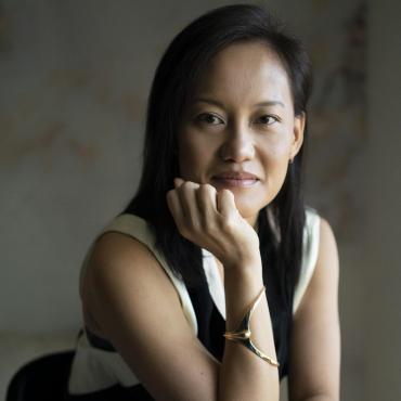 An Asian woman filmmaker smiling into the camera, resting her chin in her hand.