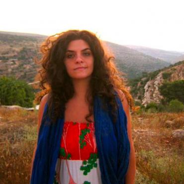 A woman with long brown hair stands in a field in front of rolling hills.