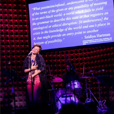 Nia Witherspoon at the microphone with a drummer and projected quote by Saidiya Hartman in the back.