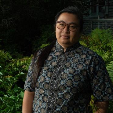 Valerie Oliveiro, mixed-race non-binary Southeast Asian in a blue, black and gray batik shirt with a mandarin collar. Surrounded by summer ferns, the image is a late evening portrait. They have their hands in their pockets looking calmly at the camera.