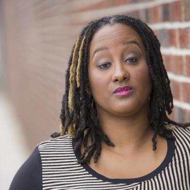 This is a headshot of Kayla Hamilton, who is a dark brown-skinned Black woman. She is posing in front of a blurred brick wall. She is wearing a long sleeve black & white striped shirt. She has light makeup and her gaze is towards us. Her black & golden highlighted dreads are down.