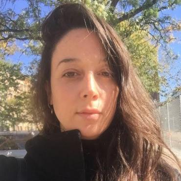 Tali Keren, a forty year old white woman with long brown hair standing against a background of trees and looking at the camera.