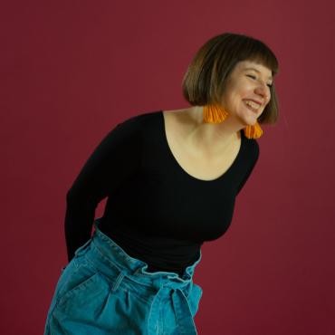 White woman with brown hair and yellow earrings leans over laughing, in front of red background.