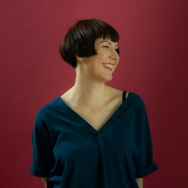 White woman with short brown hair smiles and looks to her side, in front of red background.