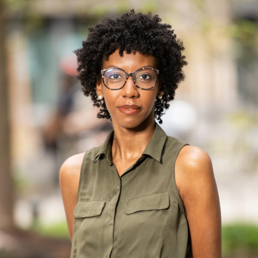 Calley N. Anderson, a 30-something, glasses-wearing Black woman playwright wearing an olive dress, looks directly at the camera with a hint of a smile.