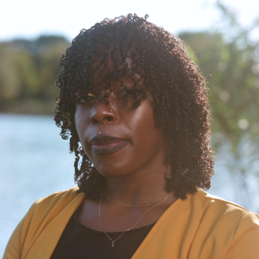 Jasmine Reid, a 30-something Black woman poet wearing a mustard yellow duster, black top, and two thin gold necklaces in front of the blurred background of a lake, looking into the camera.