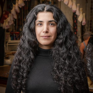 Katayoun Amjadi, a 40-something Iranian-American visual artist, standing at her studio. In the background, a sculpture of her work with hanging chicken drums frames the image.