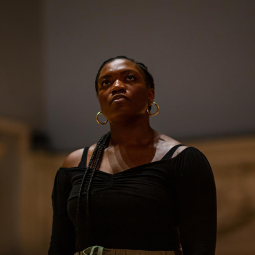 Ogemdi, a dark skinned Black woman with cornrows, stares up at the ceiling in Judson Church.