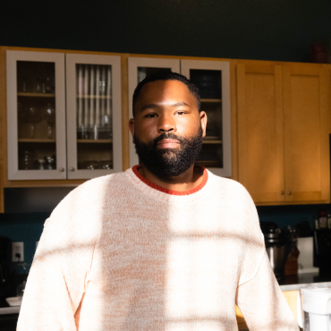 A 30-year-old Black man playwright looks into the camera with a kitchen in the back.