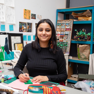 30-something woman artist sitting in her studio, wearing black, holding a pencil, looking at the camera.