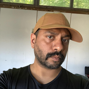 Asif Mian, a mid-40s, South Asian male artist wearing a brown leather baseball cap and black tee shirt looks intently into the camera while in his sculpture studio.