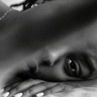 A black-and-white photo of sadé powell, a Black woman poet, lying down and gazing into the camera, with her finger partially obscuring the lens.