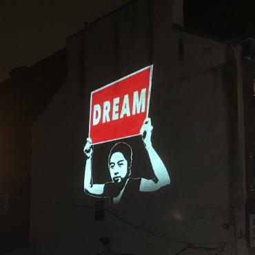 A projection on a building showing someone holding a red sign that says "Dream."